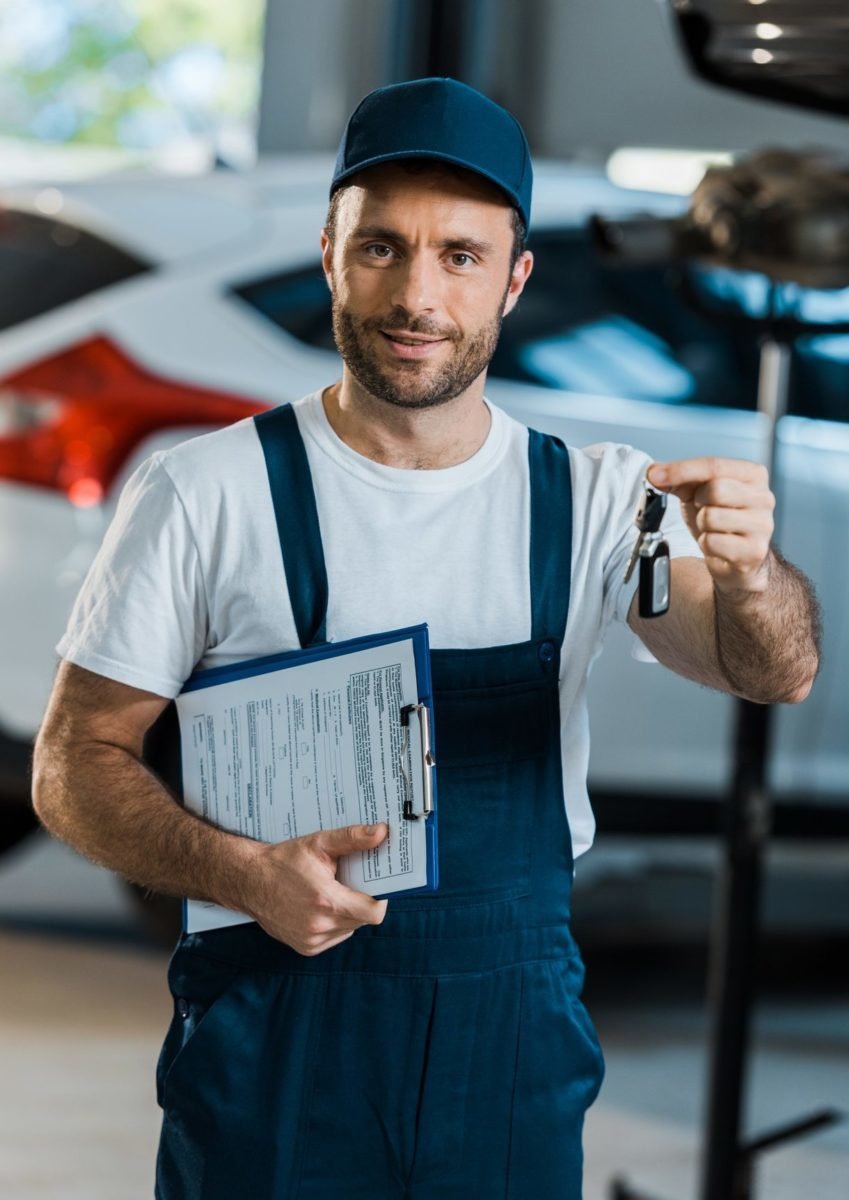 Happy Car Mechanic Looking At Camera And Holding Clipboard And Key Near Car E1651031307472